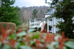 Un grand immeuble à plusieurs étages, à la façade blanche et au toit gris, entouré d’une végétation luxuriante et d’arbres.