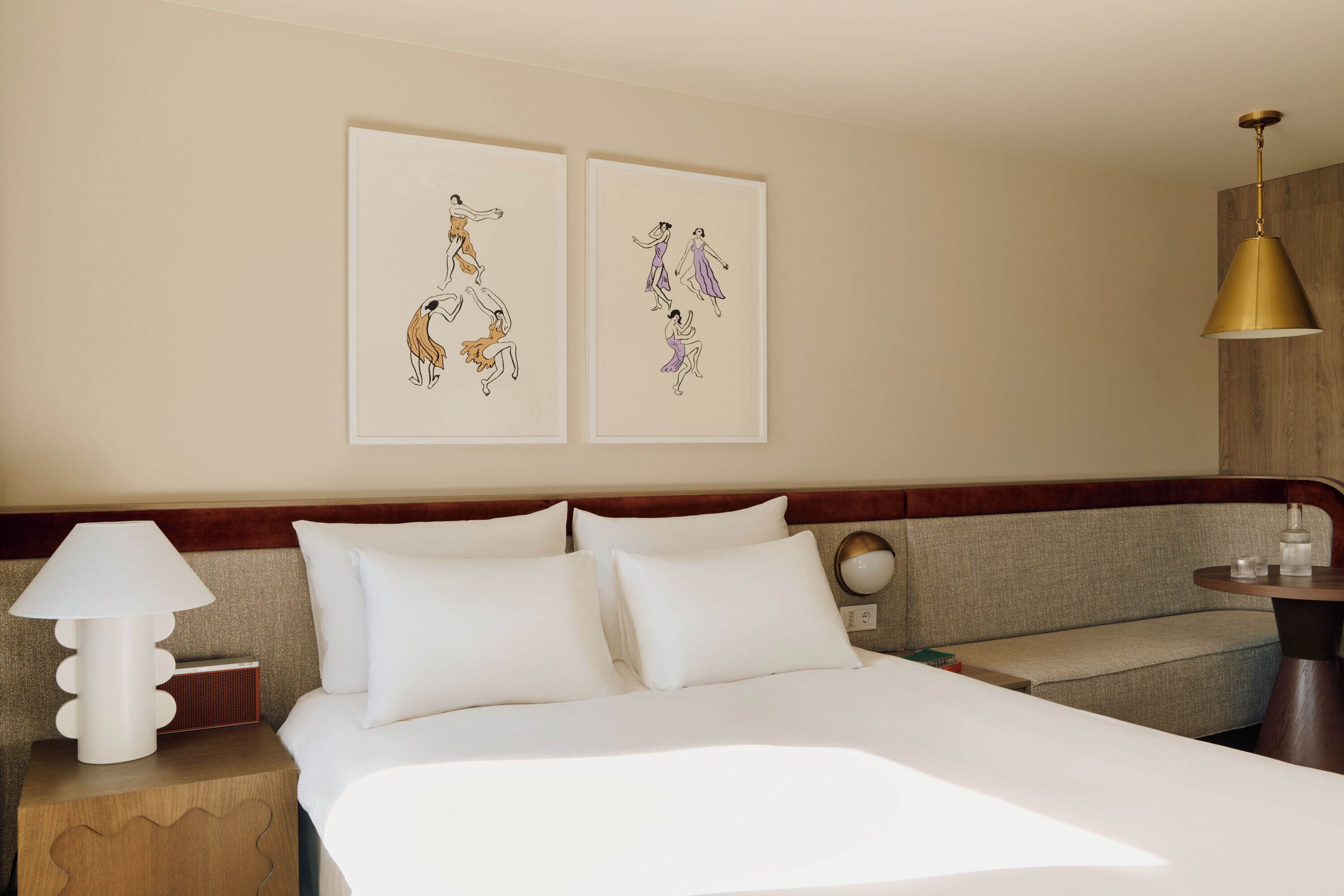 A hotel room with a bed, a nightstand with a lamp, a bedside table, and two framed artworks on the wall.