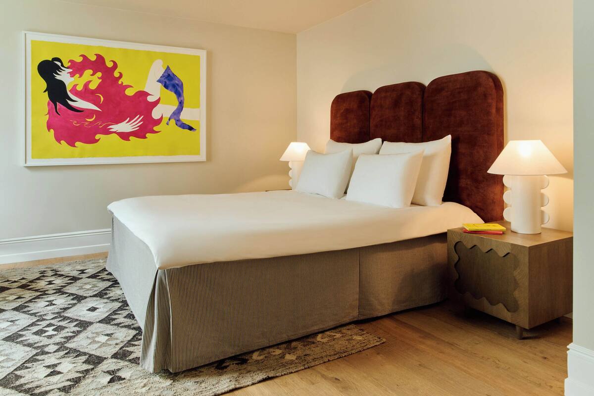 A bedroom with a large bed, two bedside lamps, a patterned rug, and a colorful abstract painting on the wall.