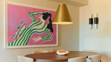A dining area with a round table, white chairs, a gold pendant light, and a framed abstract painting on the wall.