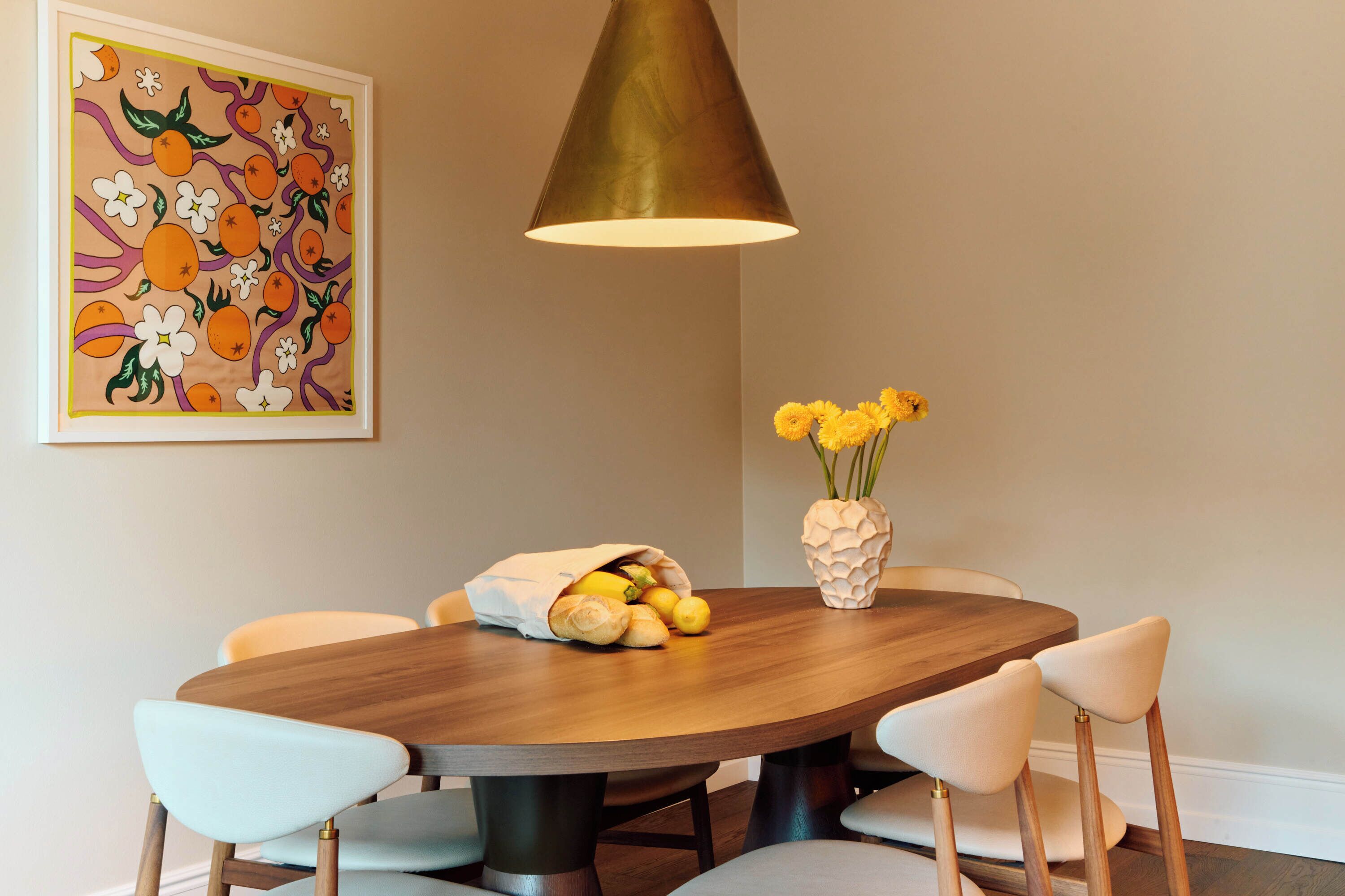 A dining area with a round wooden table, four chairs, a vase with yellow flowers, and a painting on the wall.