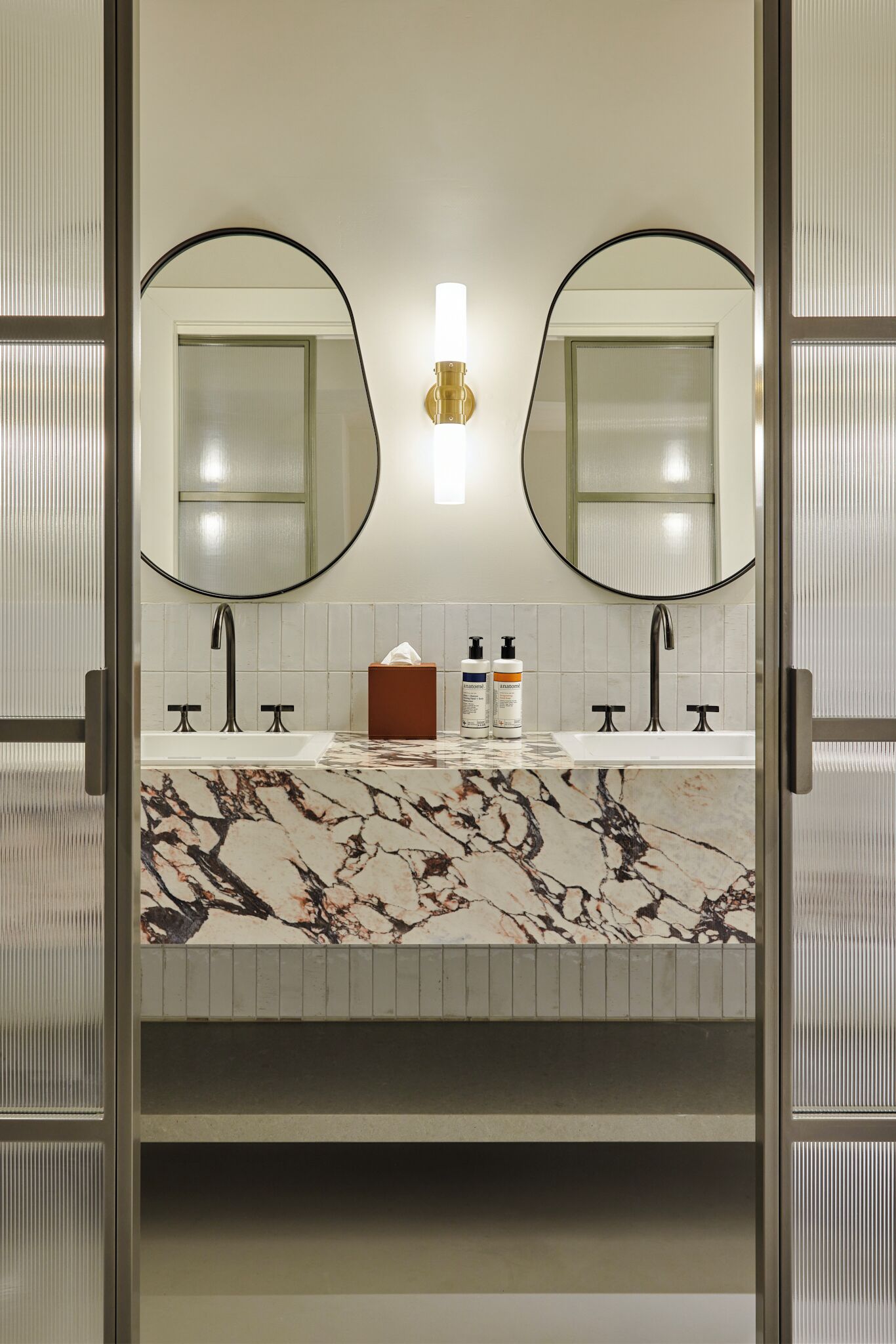 A bathroom with a marble countertop, two sinks, a round mirror, and a wall-mounted light fixture.