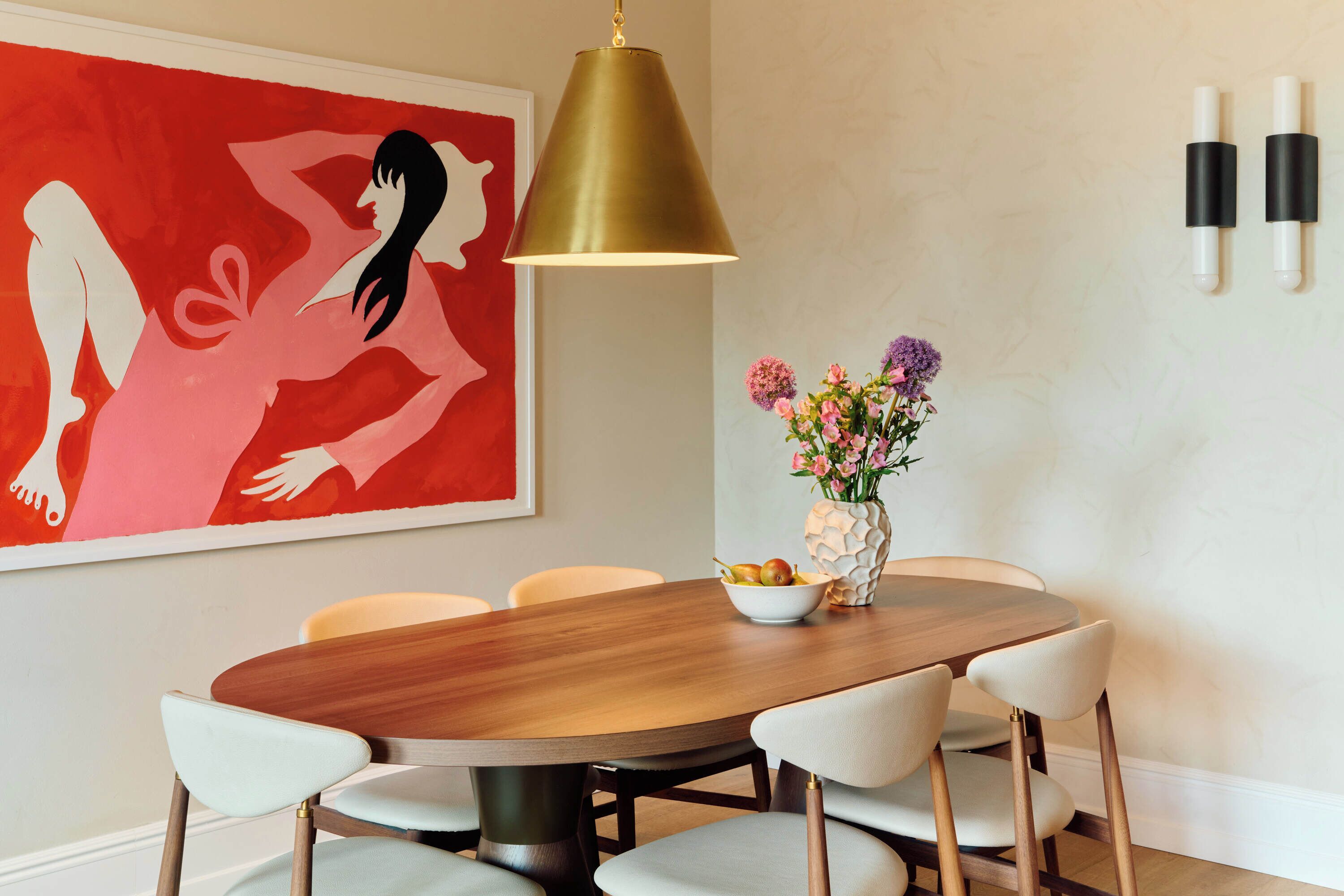 A dining area with a round wooden table, six white chairs, a vase with flowers, and a red abstract painting on the wall.