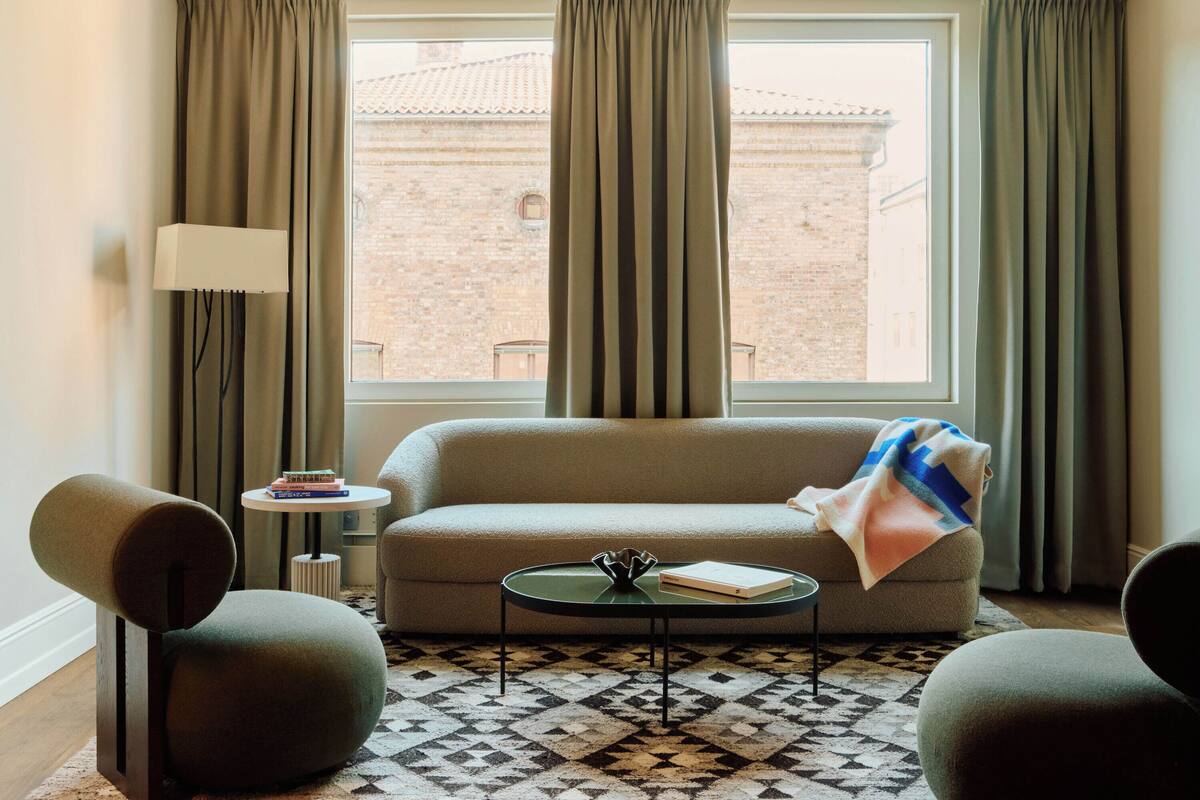 A living room with a sofa, two armchairs, a coffee table, a lamp, and a view of a brick building through a window with curtains.