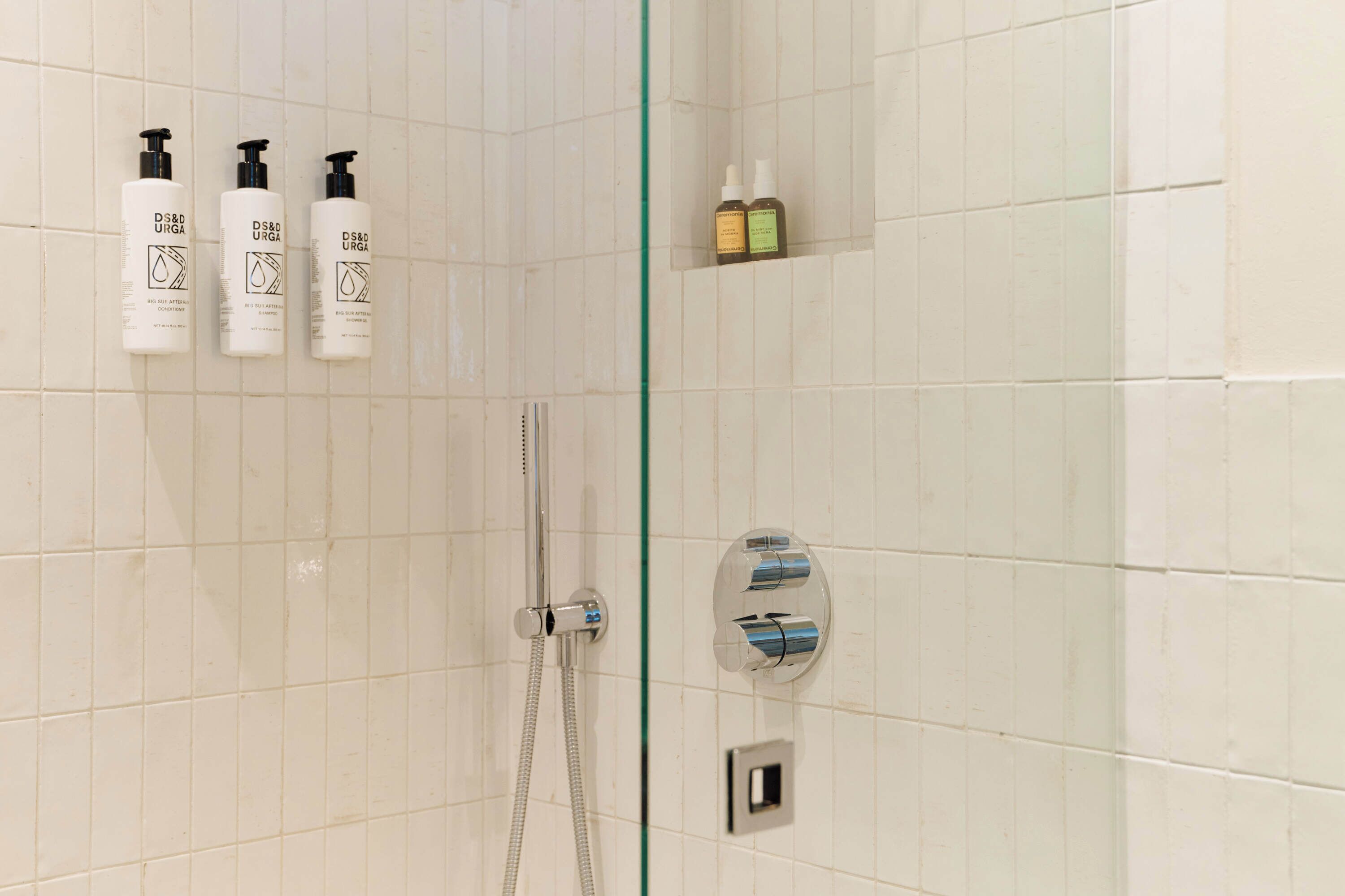 Une salle de bain aux murs recouverts de carreaux blancs, une porte de douche en verre et trois bouteilles d’après-shampoing D&S URGA accrochées au mur.