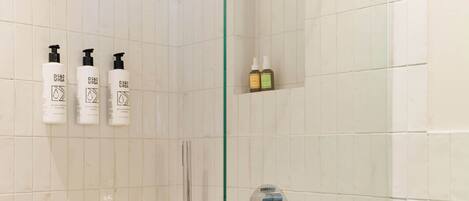 Une salle de bain aux murs recouverts de carreaux blancs, une porte de douche en verre et trois bouteilles d’après-shampoing D&S URGA accrochées au mur.