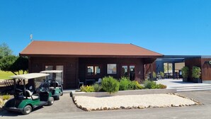 A modern building with a brown roof and wooden facade, a parking area with golf carts, and a landscaped garden with shrubs and a gravel pathway.