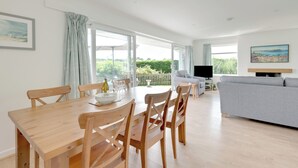 A spacious living area with a dining table set for four, a grey sofa, and a television on a stand.