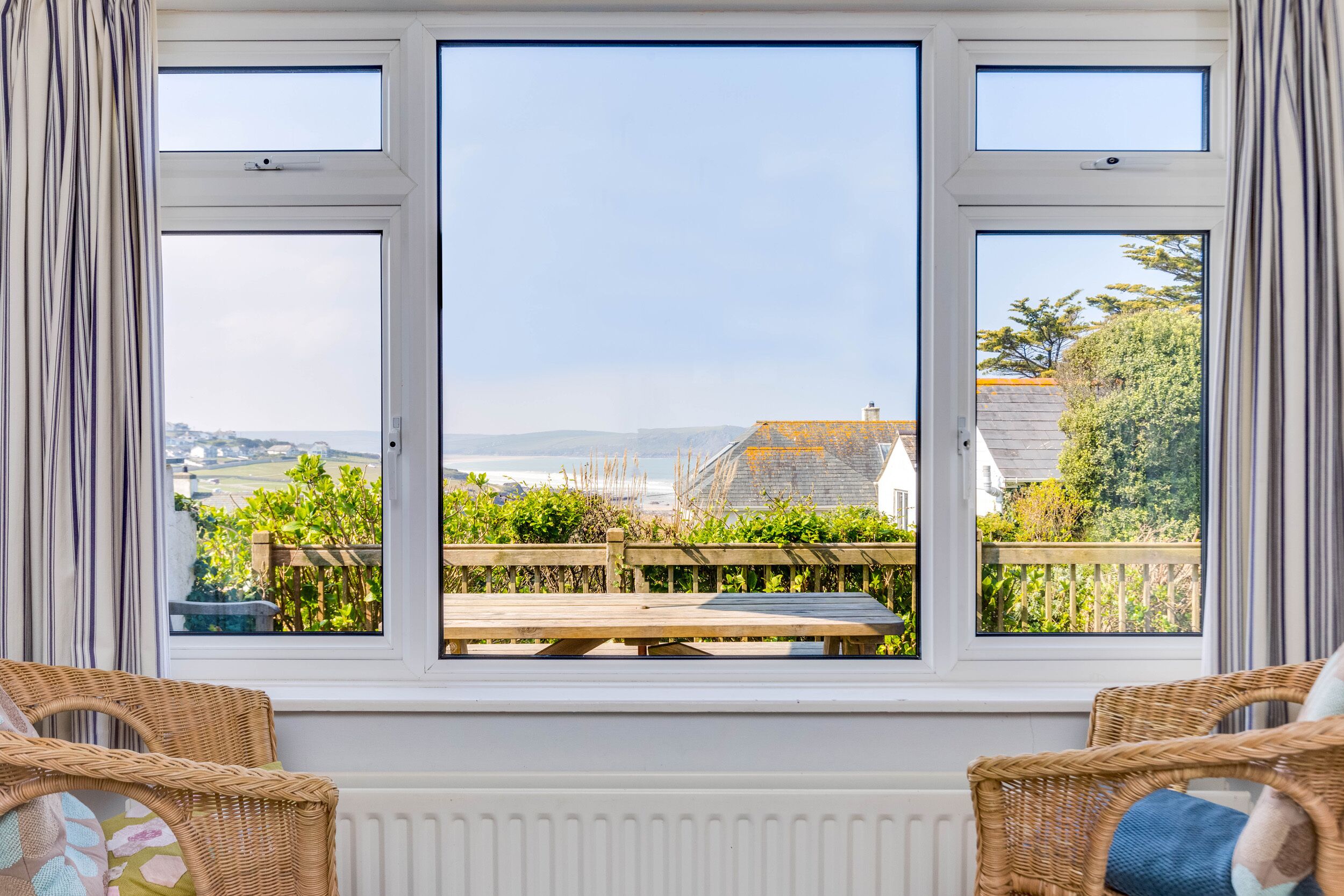 Una habitación con un ventanal grande que da a un jardín y vistas al mar. Hay sillones de mimbre y una mesa en la habitación.
