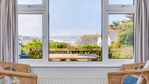 Ein Zimmer mit großem Fenster, das einen Garten und Meerblick bietet. Im Raum befinden sich Korbsessel und ein Tisch.