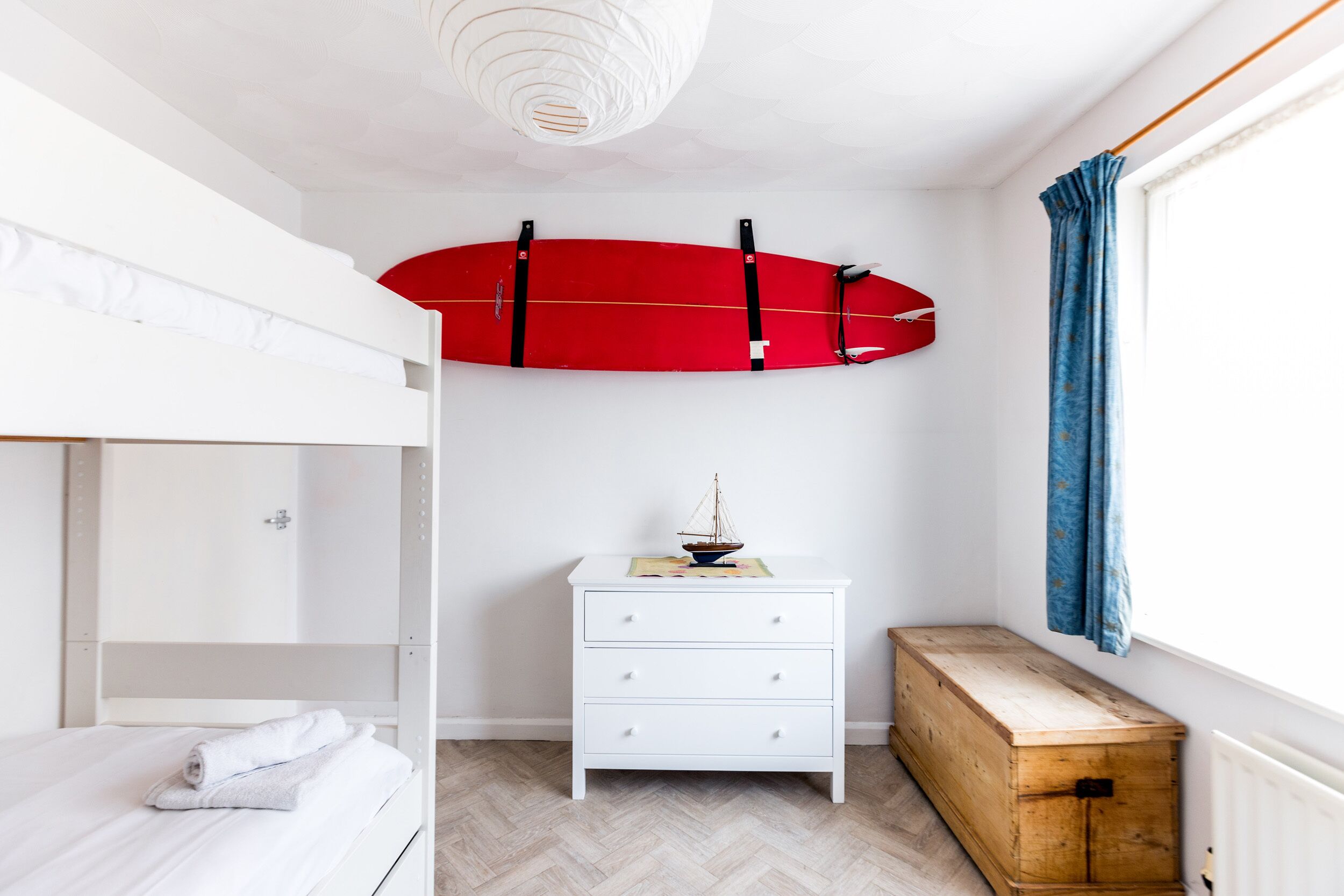 A room with a bunk bed, a white dresser, a wooden chest, and a red surfboard mounted on the wall.