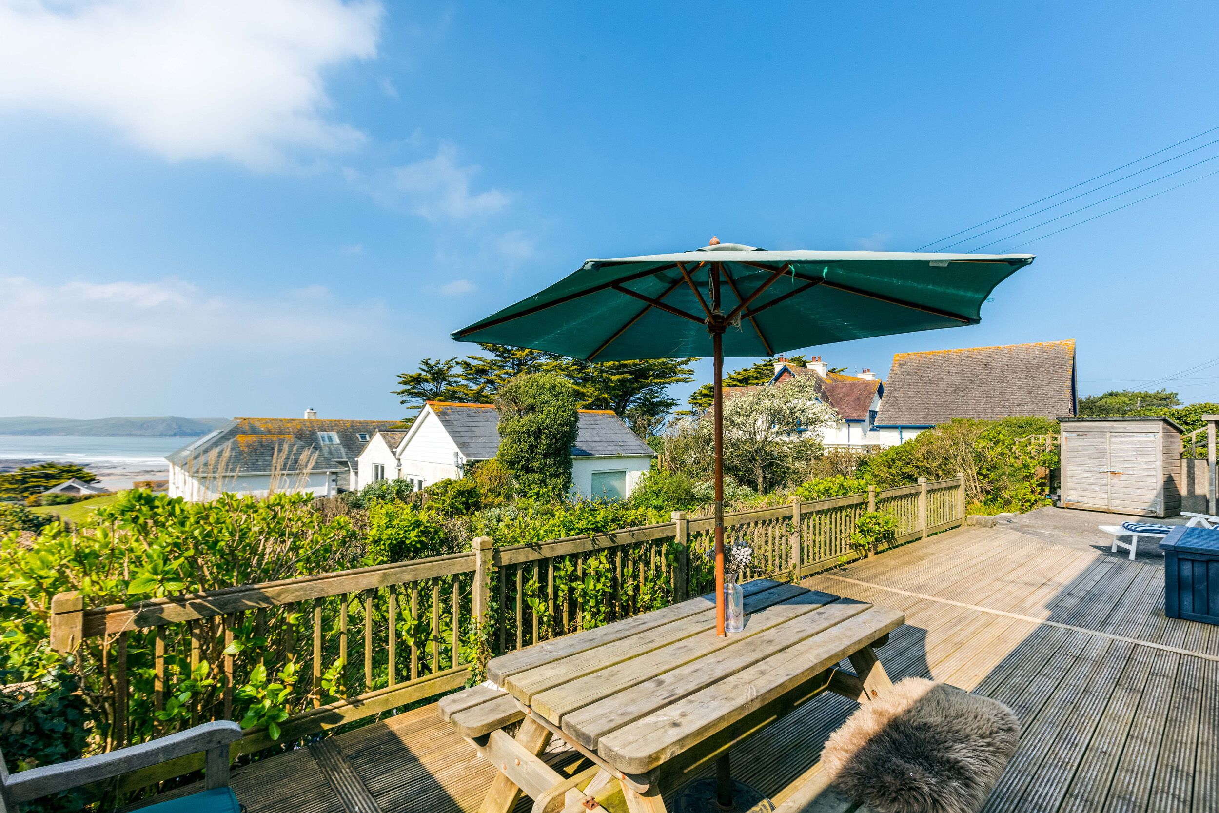 A wooden deck with a picnic table and umbrella, overlooking a coastal area with houses and greenery.