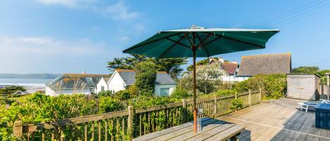 A wooden deck with a picnic table and umbrella, overlooking a coastal area with houses and greenery.