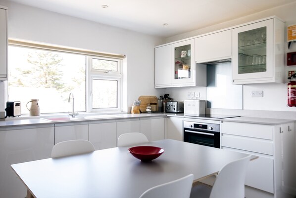 A modern kitchen with white cabinets, a dining table with chairs, a red bowl, and a window with blinds.
