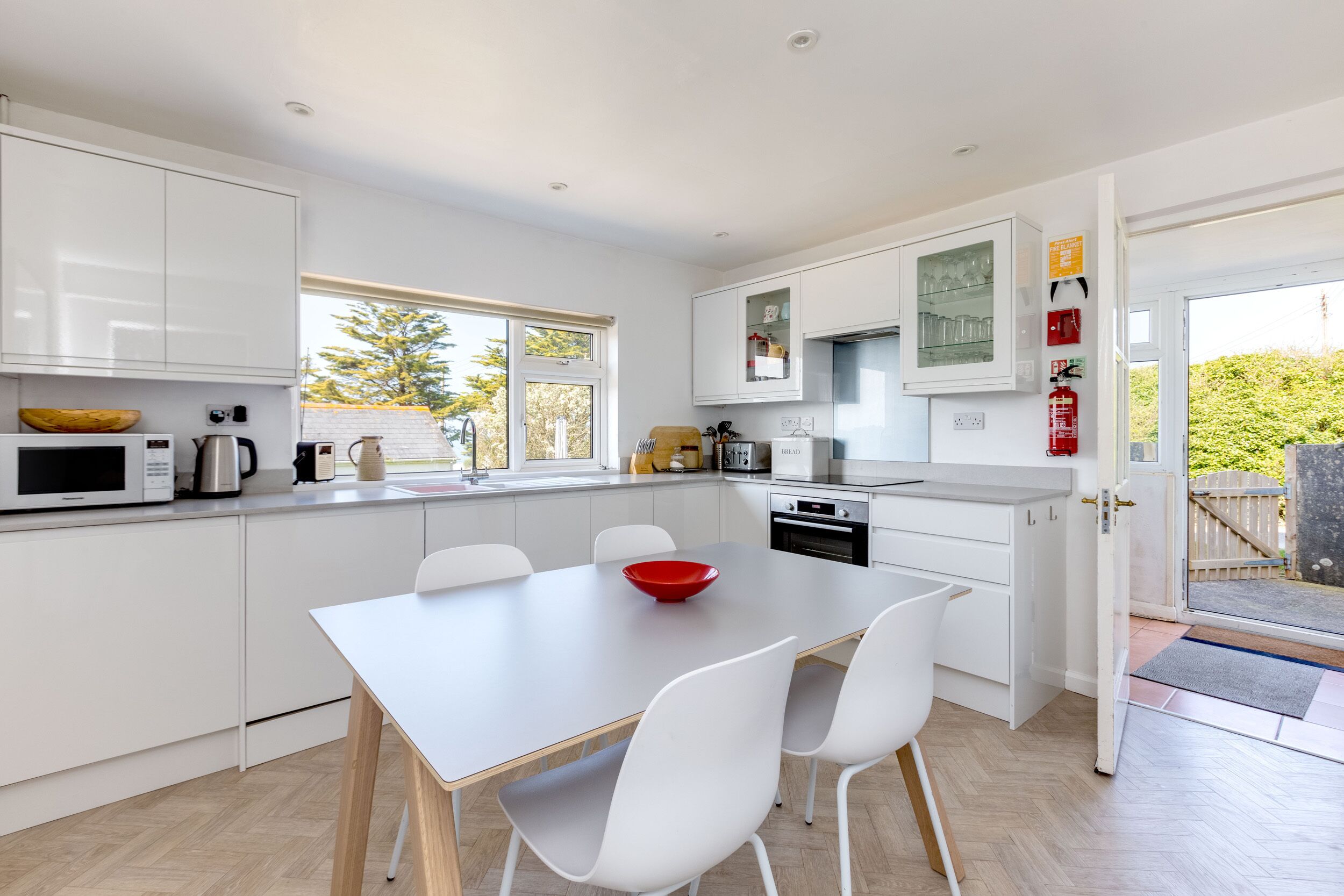 A modern kitchen with white cabinetry, a dining table with chairs, a red bowl on the table, and a view of the outdoors through the window.