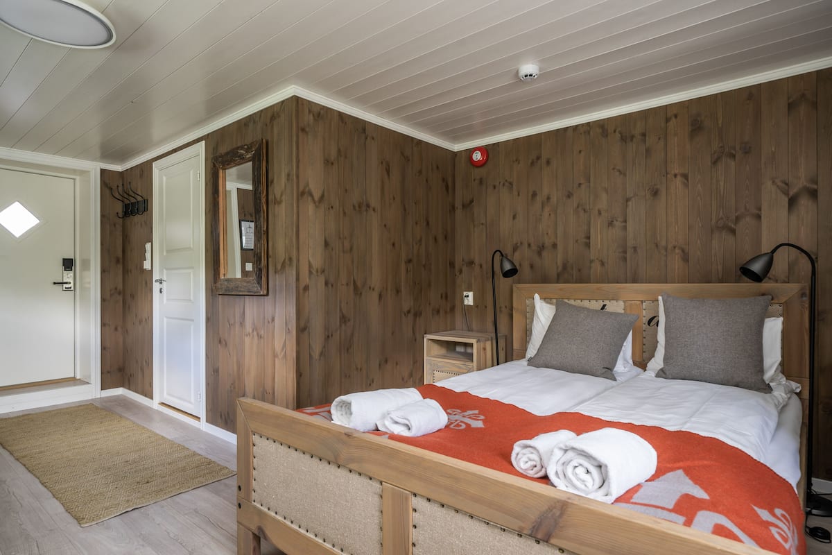 Ein Schlafzimmer mit einem hölzernen Kopfstück, einem Bett mit weißer und grauer Bettwäsche, einer roten Decke und einem kleinen Nachttisch mit Lampe.