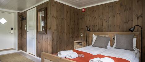Un dormitorio con cabecera de madera, cama con ropa de cama blanca y gris, una manta roja y una mesita de noche con una lámpara.