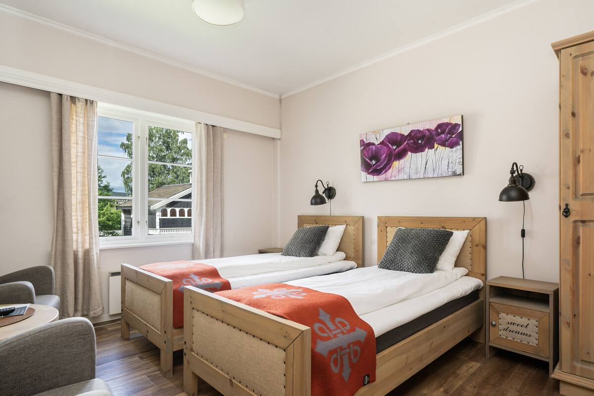 Ein Zimmer mit zwei Holzbetten, einem Schreibtisch, einem Stuhl, einem Vorhangfenster, einem Gemälde und einem Schrank.