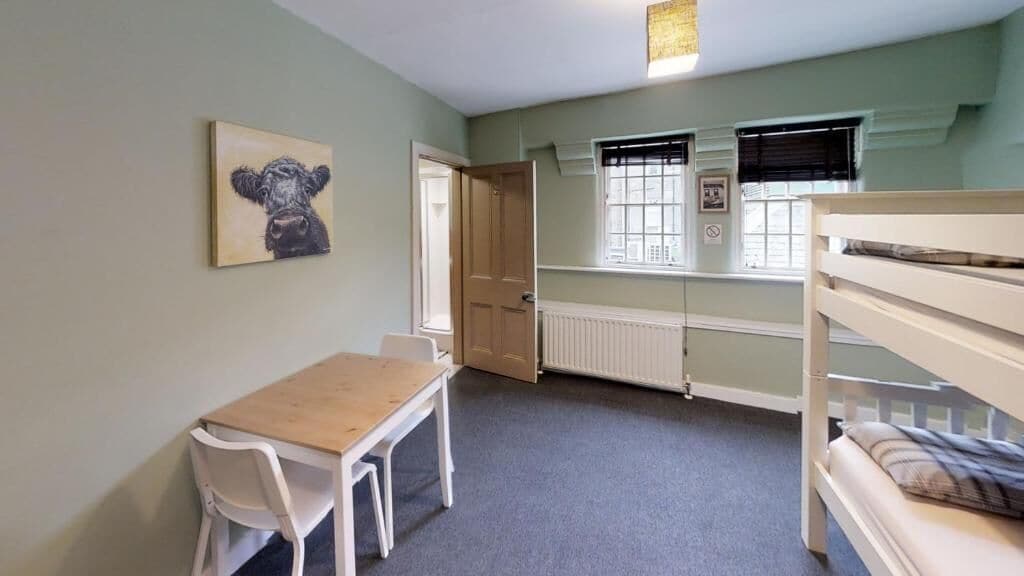 A room with a bunk bed, a table with a chair, a radiator, and a window with blinds.
