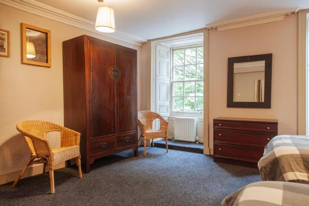 Un dormitorio con un armario grande de madera, dos sillas de mimbre, una cama con manta a cuadros, un aparador y una ventana con vistas a la vegetación.