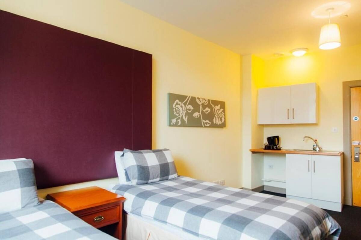Quarto de hotel com duas camas, uma kitchenette pequena e uma única obra de arte na parede.
