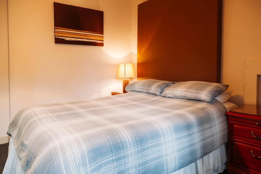 A hotel room with a bed, a nightstand, a lamp, a red dresser, and a wall with a painting.