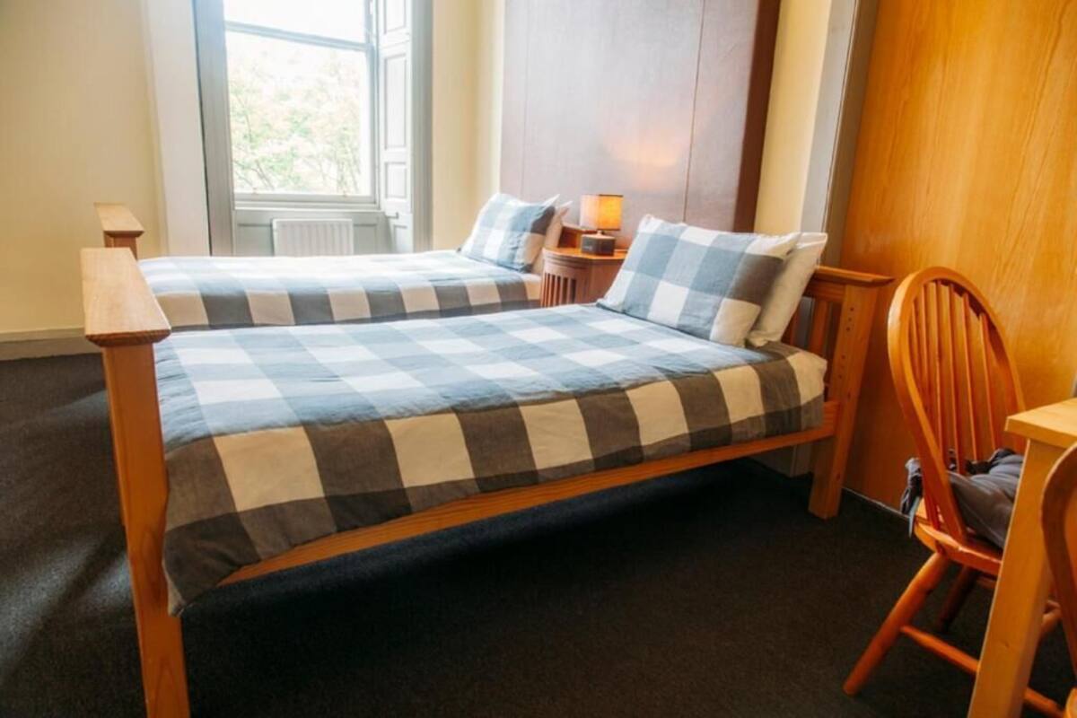 Quarto com duas camas, uma cadeira de madeira, uma mesinha e uma janela com vista para árvores.