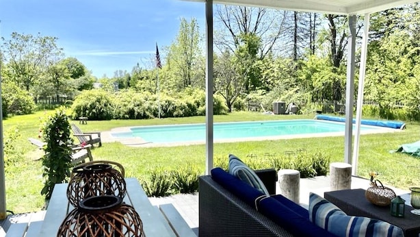A covered patio with a view of a swimming pool, surrounded by lush greenery and a wooden deck. - Spacious Adirondacks Retreat • 6BR, Heated Pool. Lake Champlain  (Willsboro)