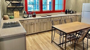A modern kitchen with wooden cabinets, a red-framed window, a built-in stove, and a dining area with a wooden table and chairs. - Spacious Adirondacks Retreat • 6BR, Heated Pool. Lake Champlain  (Willsboro)