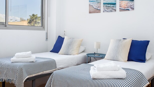 Une chambre d’hôtel avec deux lits, chacun doté de draps à rayures et d’oreillers bleus, une table de chevet avec une lampe, et une fenêtre donnant sur la plage et les palmiers.