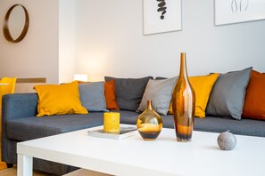 A modern living room with a white coffee table, a blue sofa with orange and gray cushions, and a large vase.