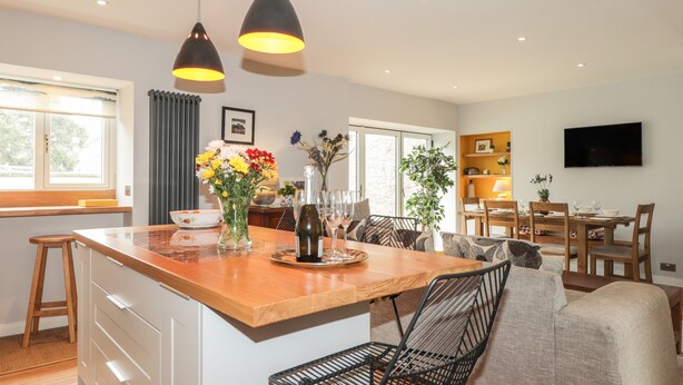 A modern kitchen with a central island, pendant lighting, and a dining area with a TV. - Old Manse (Muir of Ord)