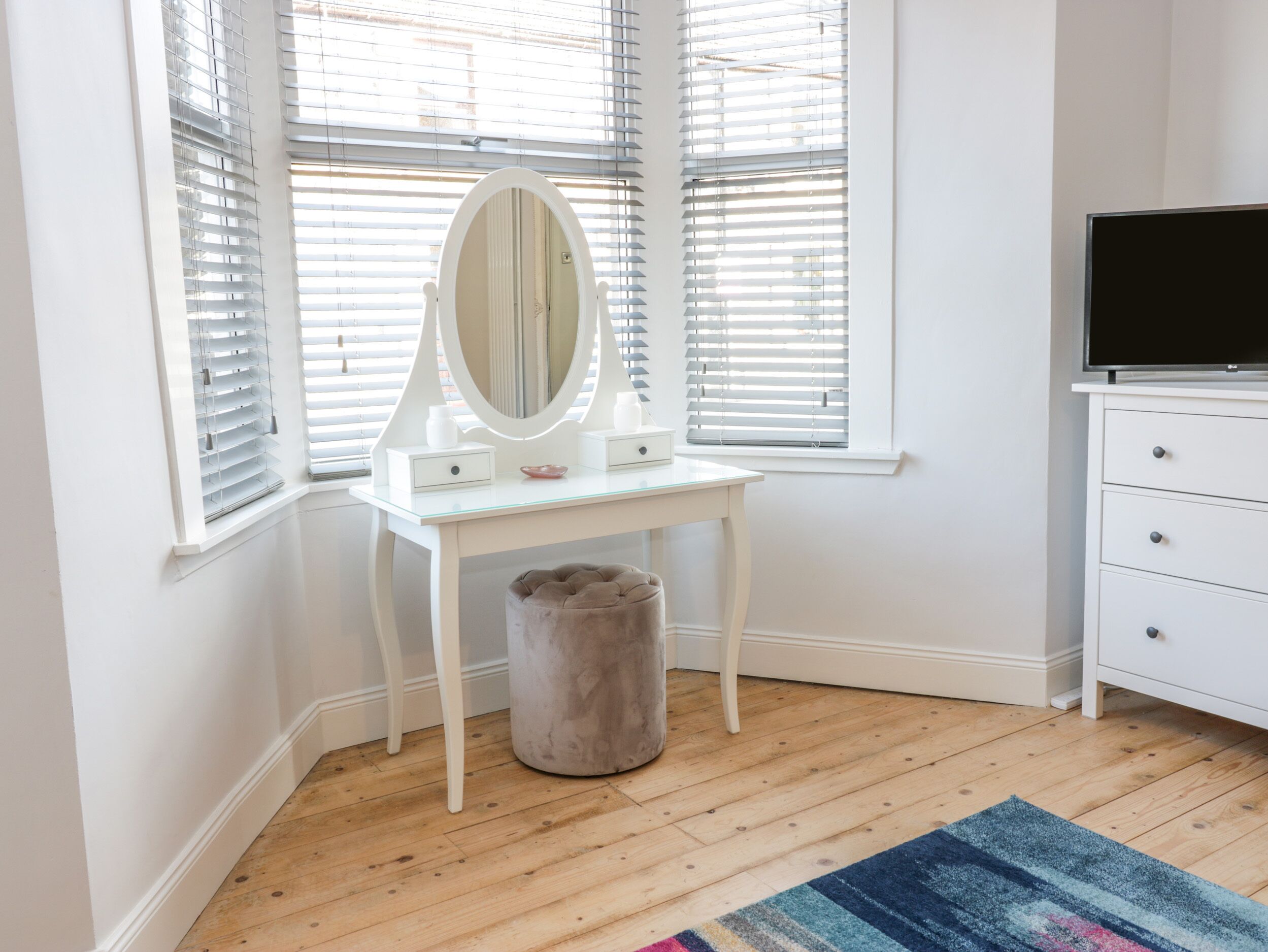 A white vanity with a mirror, a small stool, and a TV on a dresser in a room with wooden flooring and white walls.