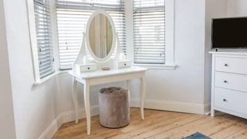 A white vanity with a mirror, a small stool, and a TV on a dresser in a room with wooden flooring and white walls.
