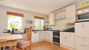 A kitchen with wooden flooring, white cabinets, a dining table with chairs, and a window with blinds. - Sandy Lodge Rock (Wadebridge)