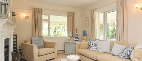 A cozy living room with a fireplace, two sofas, a patterned rug, and a view of greenery outside.