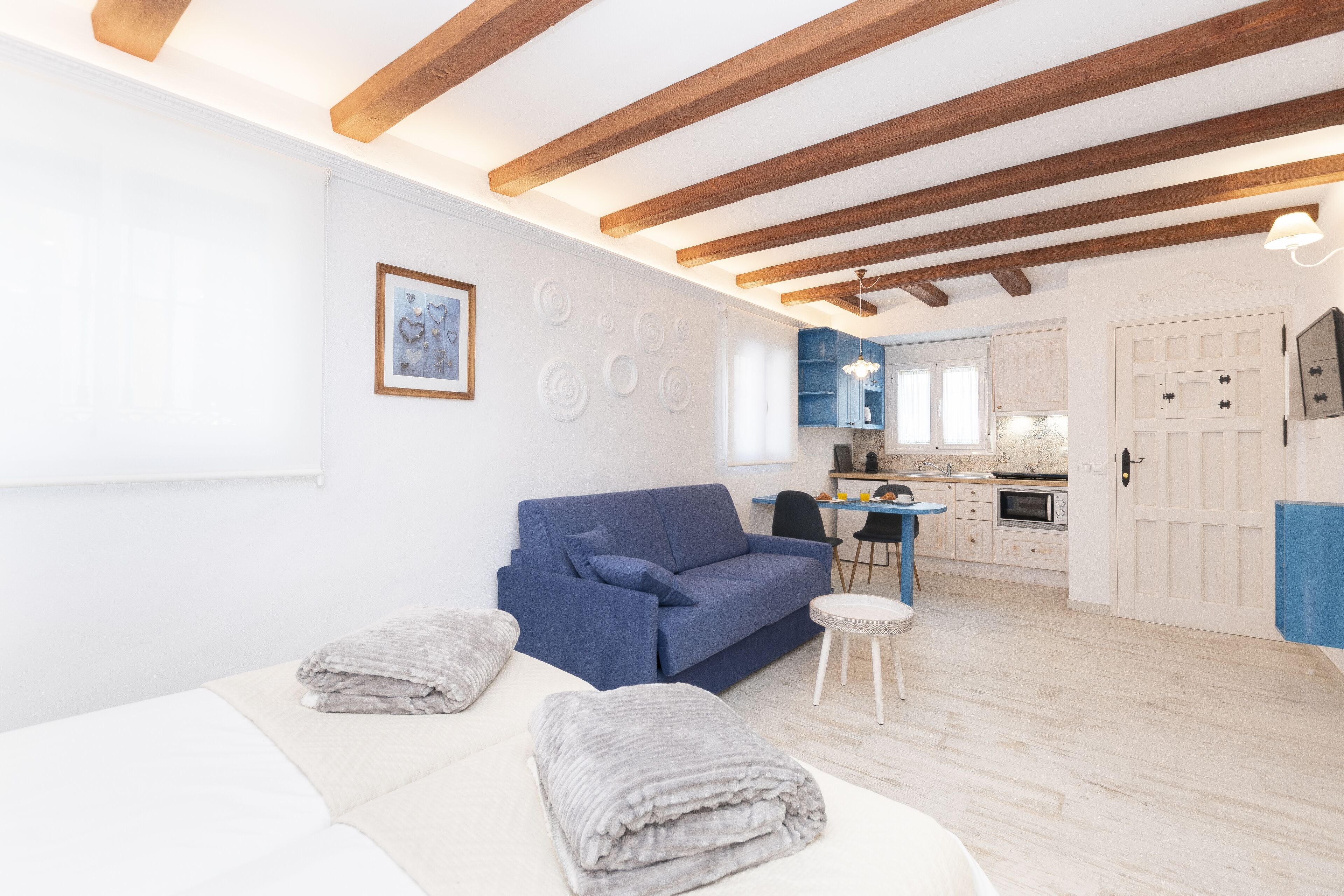 Ein modernes Wohnzimmer mit einem blauen Sofa, einem weißen Couchen und einem kleinen runden Tisch. Im Hintergrund ist eine Küche mit blauen Schränken und einer weißen Tür zu sehen.