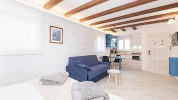Una sala moderna con un sofá azul, un sofá blanco y una pequeña mesa redonda. Se observa una cocina al fondo con armarios azules y una puerta blanca.