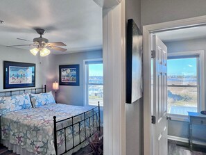 A bedroom with a bed, ceiling fan, two framed artworks, and a window with a view of buildings.