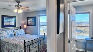 Ein Schlafzimmer mit Bett, Deckenventilator, zwei gerahmten Kunstwerken und einem Fenster mit Blick auf Gebäude.