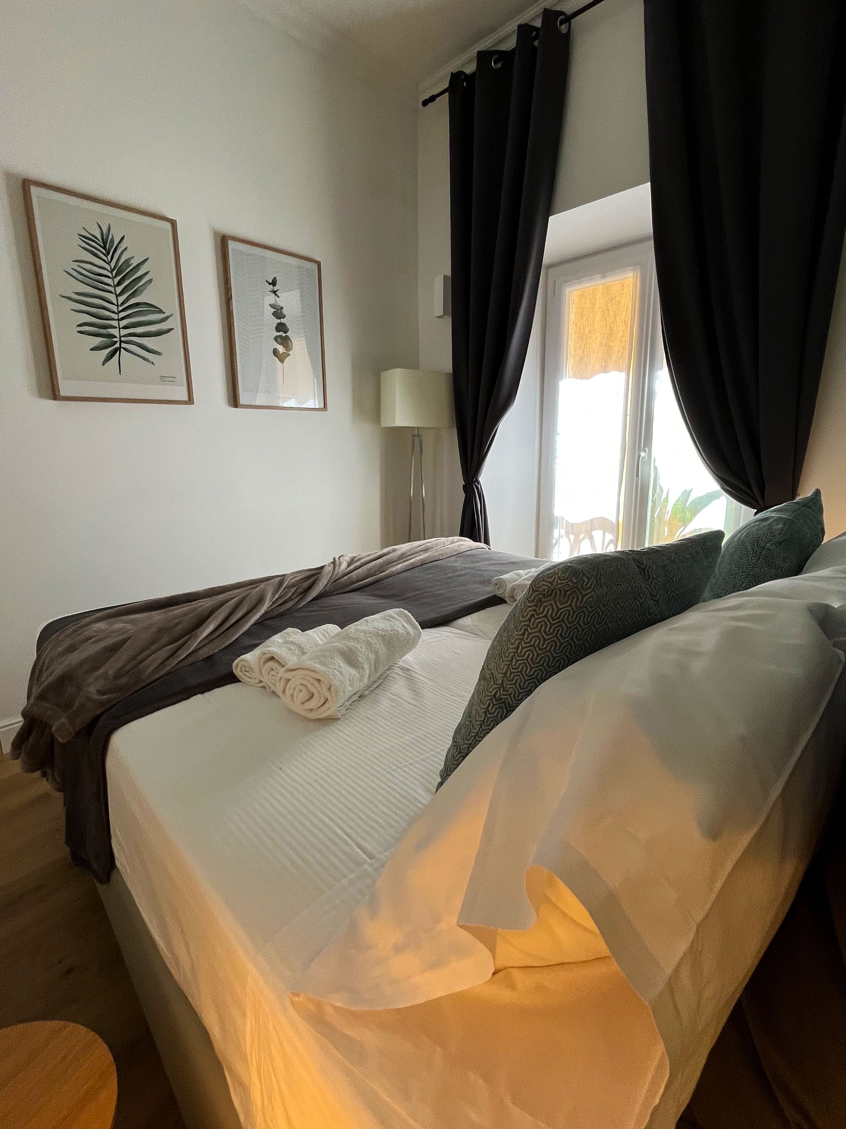A neatly made bed with a white comforter and a rolled towel, black and grey bedding, two framed botanical prints on the wall, a floor lamp, and a window with black curtains.