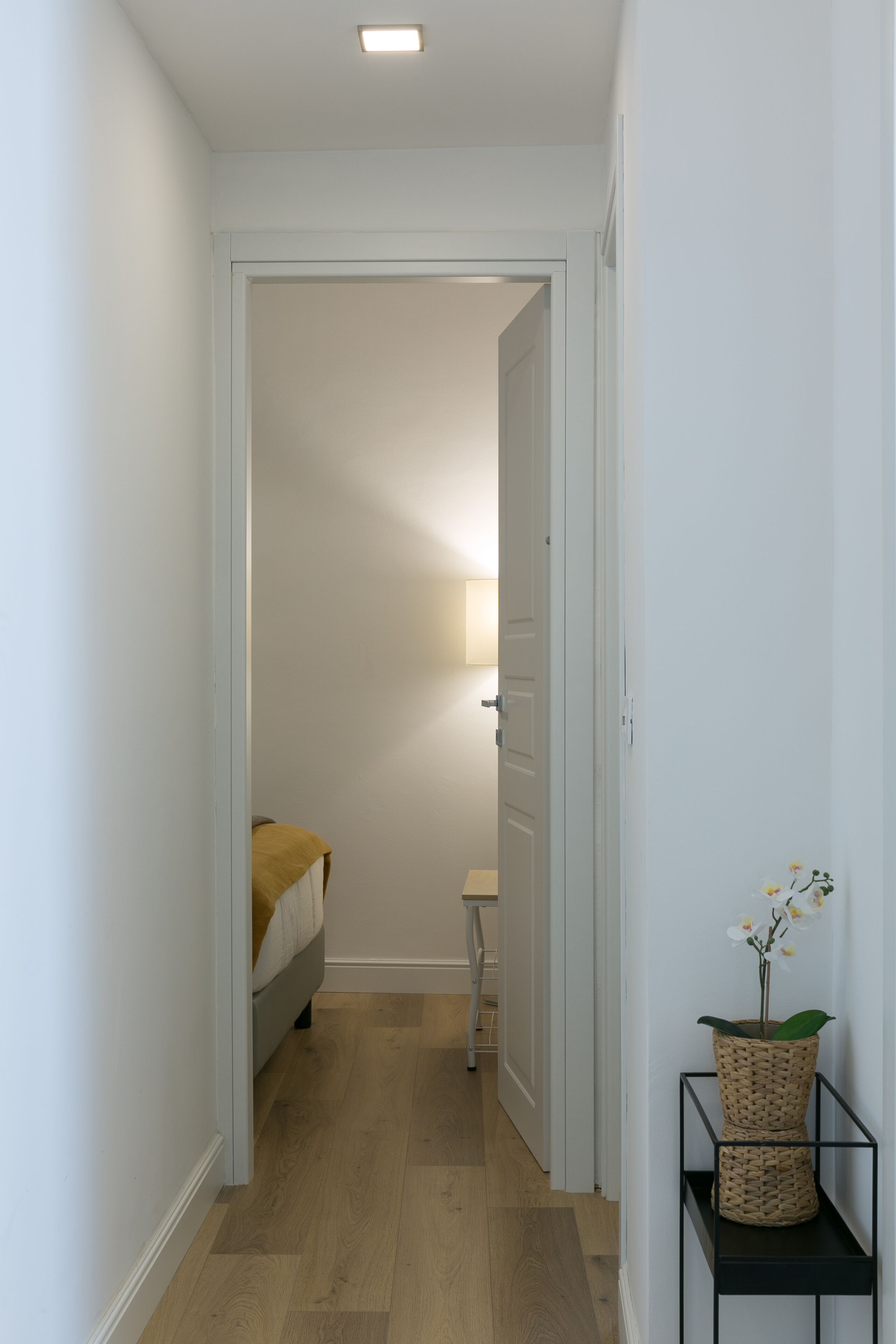 Ein schmaler Flur mit einem Bett am Ende, einer Lampe und einem kleinen Tisch mit einer Pflanze.