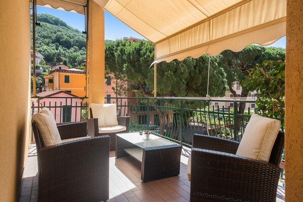 Ein Balkon mit Korbmöbeln, einem Glastisch und Blick auf eine Hügelkuppe mit Gebäuden.