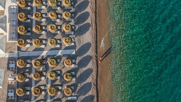 Vista aérea de un hotel frente al mar con hileras de tumbonas y sombrillas, con una vista despejada del océano y un paseo a lo largo de la playa.