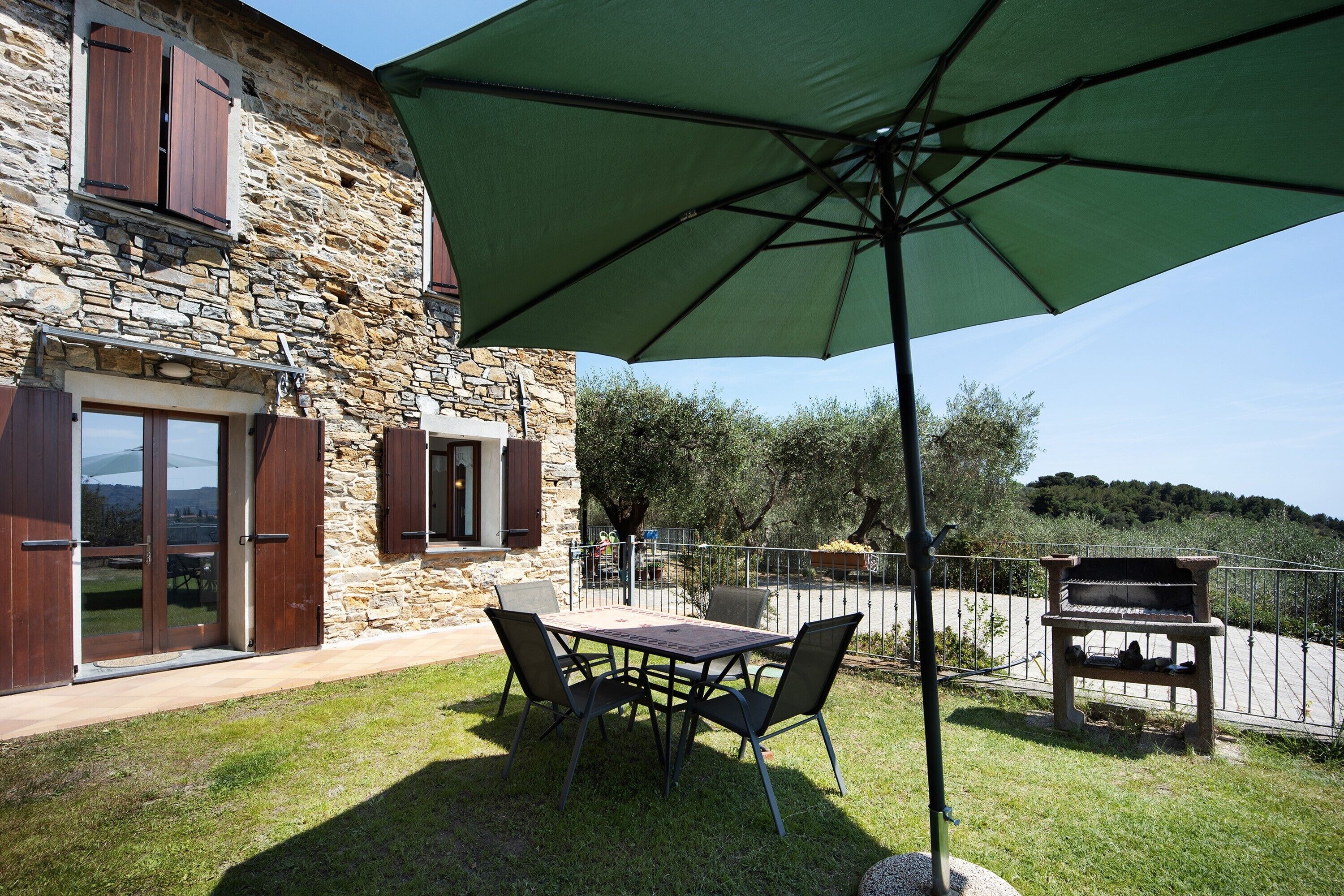 Ein Steinhaus mit einer Terrasse, einem Tisch, Stühlen, einem grünen Sonnenschirm und einem Grill.