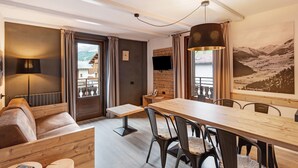 Ein modernes Zimmer mit einem Holzesstisch, schwarzen Stühlen, einer Couch, einem Fernseher und einem großen Gemälde mit Bergblick.