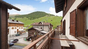 Un balcon donnant sur une montagne, une maison en bois et une rue bordée de voitures garées.