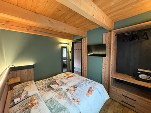 Een slaapkamer met een houten plafond, een bed met een beddeken met hertenmotief, een tv aan de muur en een houten kledingkast.