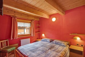 A bedroom with a bed, a chair, a window with curtains, a wooden ceiling, and wall-mounted lights.