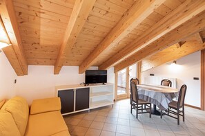 A room with a wooden ceiling, a sofa, a dining table with chairs, and a television on a cabinet.